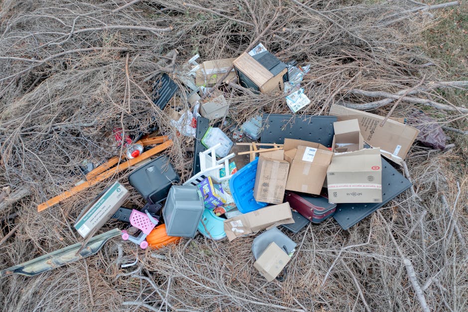 Debris comprising various discarded items such as cardboard boxes, plastic containers, a small wooden cabinet, and metal or plastic fixtures is scattered across overgrown, dried brown grasses and small twigs in an outdoor area. The waste appears to include household packaging and miscellaneous refuse, with some items stacked or leaning against each other, suggesting haphazard disposal. The scene is illuminated by natural daylight, with no visible background structures, indicating an open, neglected outdoor space possibly near a fence or boundary. This image depicts what might be cleared or removed during an independent waste collection or private rubbish removal service, highlighting the types of objects that companies like rubbishclearancewestkensington.com often handle outside traditional council collection. The clutter's arrangement and environment reflect a typical site for the collection or disposal of household or garden waste in need of professional clearance, emphasizing the importance of proper rubbish removal for such areas.