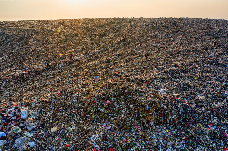 A large, expansive landfill site filled with a dense accumulation of mixed waste materials, including plastic bottles, bags, cardboard, and other household rubbish. The waste is piled high and spread unevenly across the site, with the surface displaying a combination of dark, decomposing organic matter, colorful plastic fragments, and paper debris. The textures vary from crumpled plastics to rough, layered organic material, creating a chaotic landscape of refuse. In the background, the waste continues to stretch toward the horizon under a hazy, overcast sky, with minimal vegetation or natural elements visible. The lighting appears dull and diffuse, emphasizing the scale and complexity of the waste mass. Contextually, this scene illustrates the importance of alternative waste handling solutions, such as private rubbish clearance services like those provided by [COMPANY_NAME], which endeavour to manage and reduce rubbish accumulation outside of municipal collection systems, highlighting the significance of employing professional clearance for large or difficult waste removal tasks.
