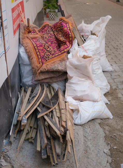 A pile of various household waste and discarded items is gathered on a pavement next to a wall. The collection includes several large, white, sealed bags filled with unidentifiable rubbish, some of which are slightly sagging or torn. Resting on top of these bags are two old, patterned rugs with intricate designs in shades of pink, purple, orange, and beige, featuring fringed edges. To the left, there are broken wooden furniture parts, such as slats and frame pieces, stacked haphazardly and appearing weathered with chipped paint and splintered edges. The scene is set outdoors in an urban environment, with a paved sidewalk and a building wall visible in the background. The lighting suggests natural daylight, and the overall setting indicates an independent rubbish removal process, possibly arranged by a private waste collection service, in line with the context of alternative rubbish clearance methods.