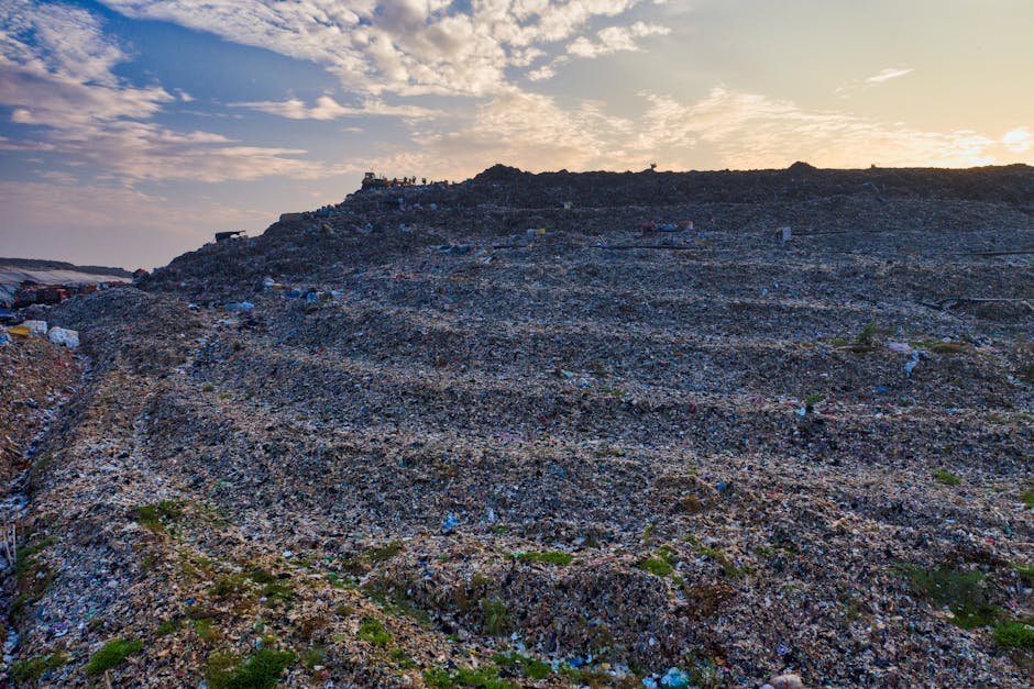 A vast landfill site extending across hilly terrain under a partly cloudy sky during late afternoon or early evening. The landscape is covered with layers of compacted waste materials, predominantly dark grey and black, with visible patches of plastic, broken furniture, and other refuse materials scattered unevenly. Small mounds of rubbish form ridges along the slopes, and the rough, textured surface shows evidence of past compaction efforts. Near the top of the hill, a small group of people is visible, standing amidst the waste, possibly observing or overseeing operations. Sparse patches of vegetation, such as small shrubs and grass, are interspersed among the debris, providing a slight contrast to the otherwise industrial scene. The environmental context suggests an unmanaged or semi-managed waste disposal area that could be accessed for independent rubbish collection or private disposal services, aligning subtly with alternative waste handling solutions. Natural lighting from the setting sun casts a warm glow over the site, enhancing the details of the waste and terrain for clarity and observational accuracy.