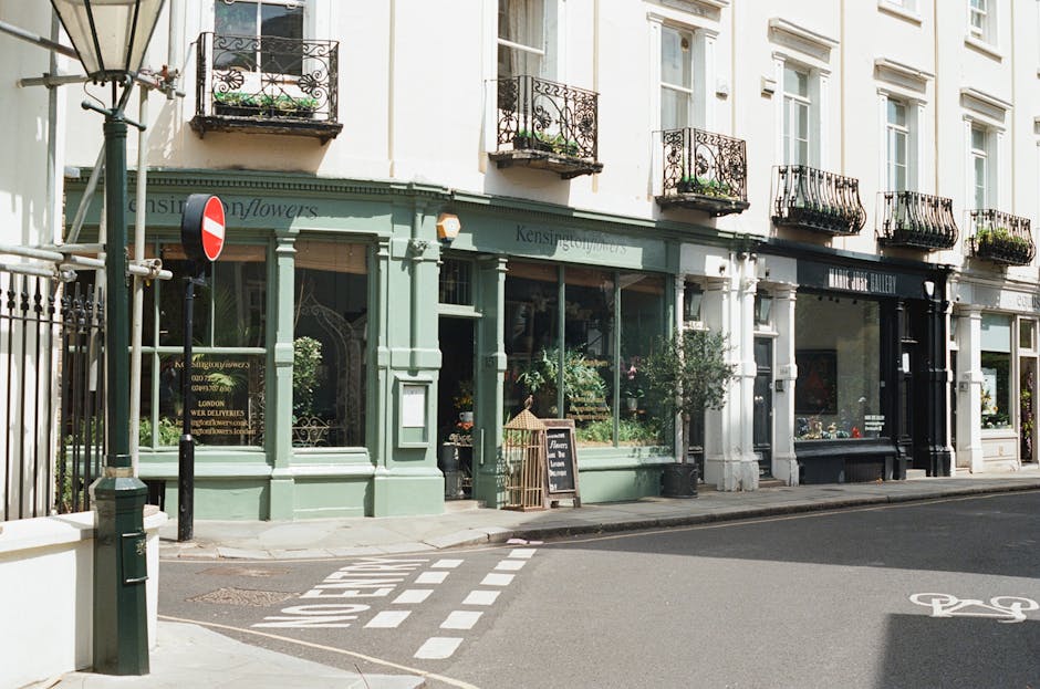 The image depicts a quiet urban street scene featuring a shop with a light green facade, situated on the ground floor of a white multi-story building with several small balconies adorned with black wrought iron railings and potted plants. The shop's large glass windows display various plants and items inside, and a blackboard sign stands outside the entrance. To the left, there is a street lamp with a traditional design, and a metal pole with a no entry traffic sign. The pavement is clean, and a designated bicycle lane with painted symbols runs along the road. The overall scene appears well-maintained and bright, with natural light illuminating the storefront and street, suggesting a typical daytime setting. This environment is relevant to waste and rubbish management, as the tidy condition and organized street infrastructure support efficient rubbish removal and private waste handling practices, such as those offered by services like rubbishclearancewestkensington.com in the West Kensington area.