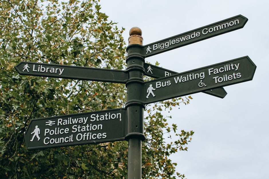 A black metal directional street signpost situated outdoors against a background of a leafy tree with green and some yellowing leaves. The signpost features multiple rectangular signs with white text and symbols, pointing in various directions. The signs indicate nearby locations including Biggleswade Common, Library, Railway Station, Police Station, Council Offices, Bus Waiting Facility, and Toilets. The sign for the railway station and police station includes a symbol of a train, while the bus waiting facility and toilets sign features a wheelchair accessible symbol and a pedestrian icon. The primary focus is on the signpost’s sturdy cylindrical pole and the clear, legible signs, reflecting an environment where clear directional guidance is essential. The lighting appears natural, suggesting daytime with overcast skies, and the overall scene emphasizes the function of providing accessible navigation in a community setting, aligning with principles of local and alternative waste and rubbish management by highlighting organized, public infrastructure.