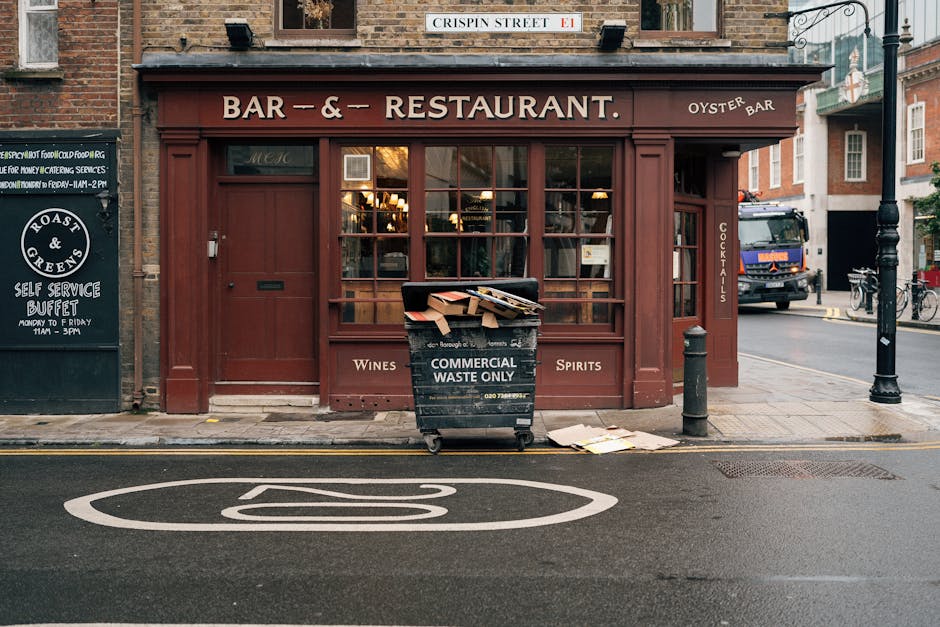 The image depicts a quiet urban street scene featuring a shop with a light green facade, situated on the ground floor of a white multi-story building with several small balconies adorned with black wrought iron railings and potted plants. The shop's large glass windows display various plants and items inside, and a blackboard sign stands outside the entrance. To the left, there is a street lamp with a traditional design, and a metal pole with a no entry traffic sign. The pavement is clean, and a designated bicycle lane with painted symbols runs along the road. The overall scene appears well-maintained and bright, with natural light illuminating the storefront and street, suggesting a typical daytime setting. This environment is relevant to waste and rubbish management, as the tidy condition and organized street infrastructure support efficient rubbish removal and private waste handling practices, such as those offered by services like rubbishclearancewestkensington.com in the West Kensington area.