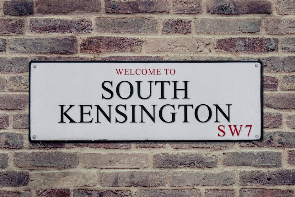 A rectangular metal street sign mounted on a red and brown brick wall displaying the words 'Welcome to South Kensington' in bold black letters. The sign has a white background with a thin black border and features the postcode 'SW7' in red at the bottom right corner. The bricks around the sign vary in shade, with some appearing more weathered and textured, providing a rustic contrast to the clean, modern design of the sign. Natural daylight illuminates the scene evenly, highlighting the detailed textures of the brick surface and the reflective surface of the sign, which is flush against the wall. This type of signage can often be seen in residential or community areas where private or independent collection alternatives for rubbish and waste disposal are common, aligning with services like rubbish clearance and on-site waste handling. The installation of this sign suggests a typical urban or suburban setting where local regulations and community identity are expressed through street signage, without visible debris or waste in the image.