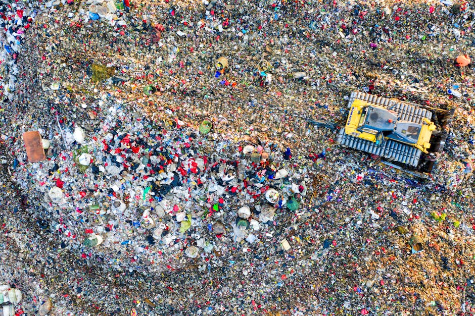 An aerial view of a landfill site showing a large expanse of compacted mixed waste, including plastics, paper, textiles, and organic debris, spread across a sloped terrain. The waste is a colorful mixture with red, white, blue, green, and other hues, with some materials partially buried under others. Near the right side of the image, a small yellow tracked excavator with a compact body and metal tracks is visible, positioned on top of the waste, likely engaged in debris movement or collection. The surrounding environment appears barren and industrial, with no natural vegetation visible, emphasizing the scale and density of the waste. The scene illustrates the process of waste management and disposal, potentially involving private waste handling services such as those provided by rubbish clearance specialists like rubbishclearancewestkensington.com, used as an alternative to traditional council rubbish collection methods. The lighting suggests daylight, and the overall atmosphere highlights an outdoor, operational landfill or waste processing area.