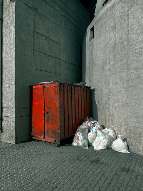 A large, rusty red metal skip positioned against a concrete wall in an outdoor urban setting. The skip appears weathered, with visible patches of rust and chipped paint, indicating wear and exposure to the elements. Surrounding the skip are several tied plastic garbage bags filled with mixed waste, including clear and black bags that are slightly misshapen and bulging, suggesting they are filled with household or construction rubbish. The skip is situated on a dark, textured paving surface, with a tall, dark-colored building wall on the left and a lighter, smooth concrete wall behind the skip on the right. In the background, part of a modern-looking structure with a small, narrow window or vent can be seen, integrating into a city environment where private or independent waste handling may be occurring. The overall scene reflects a typical scenario of on-site rubbish disposal, possibly linked to alternative waste management practices, with minimal natural light casting soft, diffuse shadows.