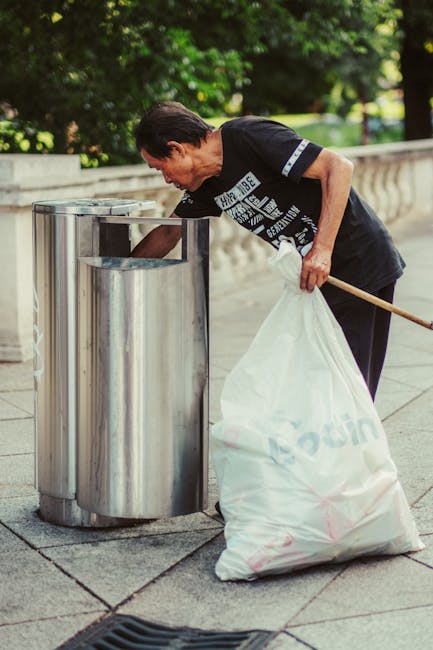 A man with dark hair, wearing a navy blue t-shirt with white and blue graphics on the front, is seen using a long-handled tool to pick up waste from a stainless steel public trash bin placed on a paved sidewalk. He is holding a large white plastic garbage bag, partially filled, in his other hand. The setting appears to be outdoors in a park or urban area, with lush green trees and a stone or concrete railing in the background. The trash bin is cylindrical with a smooth, shiny surface, and is situated alongside other similar bins, aligned in a row. The lighting is natural, casting soft shadows, and the overall scene depicts a routine public waste collection effort, which might relate in context to private rubbish clearance services like those provided by rubbishclearancewestkensington.com, especially for on-site waste removal or alternative disposal methods. The environment reflects a clean, well-maintained public space, with the man actively engaged in rubbish collection or waste management task, reinforcing the importance of proper waste disposal in urban areas.