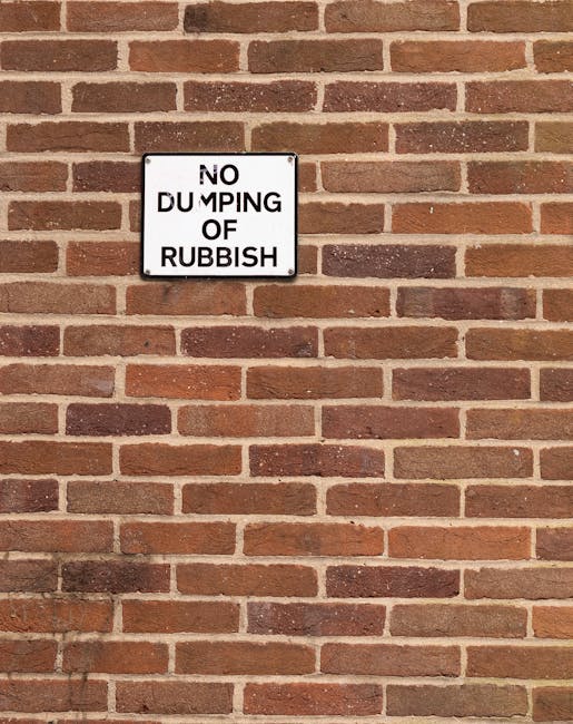 A large, rusty red metal skip positioned against a concrete wall in an outdoor urban setting. The skip appears weathered, with visible patches of rust and chipped paint, indicating wear and exposure to the elements. Surrounding the skip are several tied plastic garbage bags filled with mixed waste, including clear and black bags that are slightly misshapen and bulging, suggesting they are filled with household or construction rubbish. The skip is situated on a dark, textured paving surface, with a tall, dark-colored building wall on the left and a lighter, smooth concrete wall behind the skip on the right. In the background, part of a modern-looking structure with a small, narrow window or vent can be seen, integrating into a city environment where private or independent waste handling may be occurring. The overall scene reflects a typical scenario of on-site rubbish disposal, possibly linked to alternative waste management practices, with minimal natural light casting soft, diffuse shadows.