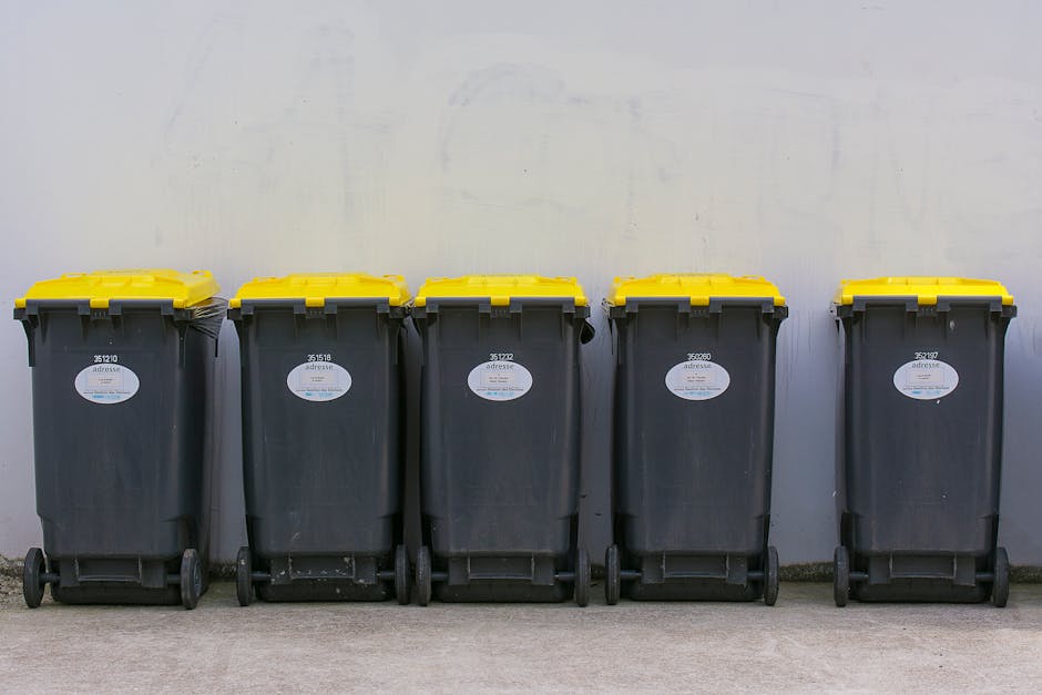 Five large wheeled rubbish bins with dark grey bodies and bright yellow lids are lined up against a plain, light grey wall in an outdoor setting. The bins are positioned on a concrete surface, with each bin's front facing forward, and they are evenly spaced, with their wheels visible at the bottom. The bins have white labels with barcode markings and small printed text, possibly indicating disposal codes or identification numbers. The scene is illuminated by natural daylight, casting soft shadows on the ground, and the background wall appears smooth and unadorned. This setup suggests a private or commercial waste collection area where independent disposal methods, such as these bins, are used for rubbish removal services, aligning with practices in on-site clearance or alternative waste handling outside of local authority collection systems, as seen in the context of rubbish clearance services in West Kensington.