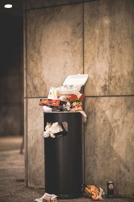 Outside on a paved pavement in front of a green metal fence with decorative white graffiti-style artwork, there is a collection of discarded cardboard boxes and packaging materials, including an open purple fruit box, a red plastic crate, and a black box with gold and white details. A concrete trash bin, placed on a designated base, contains red foam packaging material. Surrounding the bin are additional packaging waste and small plastic remnants. The environment appears to be an urban outdoor setting, with the clutter indicating waste awaiting collection or disposal. The scene reflects a typical area where private or independent rubbish removal services, such as those provided by companies like rubbishclearancewestkensington.com, might manage the collection of general waste and packaging debris, supporting proactive rubbish management outside communal recycling or municipal waste systems.