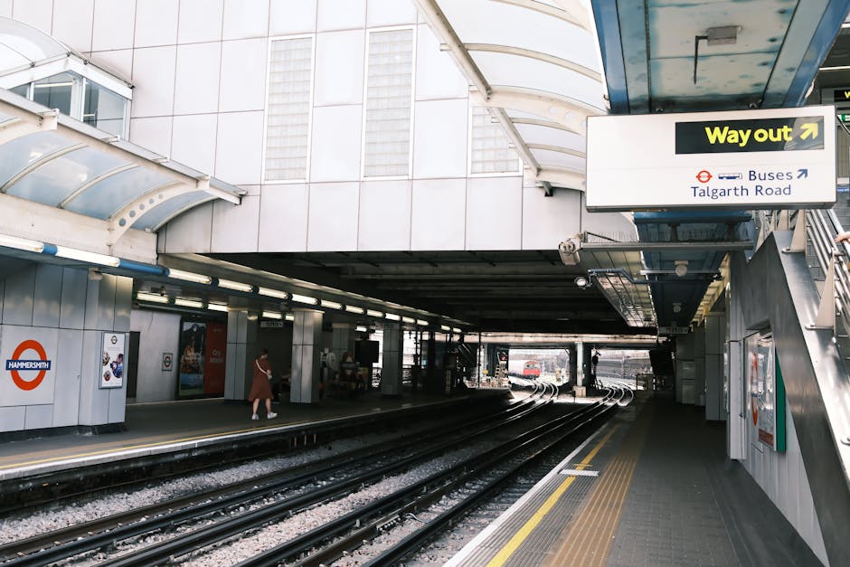 At a modern train station platform, a worker dressed in dark clothing and a beanie, wearing a high-visibility vest, stands beside a large hand cart loaded with a variety of waste bags and discarded items, including black, grey, and orange rubbish bags, some crumpled or tied, and a small cardboard box. Behind the worker, there are mechanical lifting devices and safety equipment mounted on a glass-walled structure, indicating a facility equipped for waste handling or industrial cleanup. To the left, a white recycling bin is visible on the tiled platform surface. The train tracks run parallel along the platform, with a passing train partially visible on the right, and additional platform signage and distant figures can be seen further down the station. The overall scene is illuminated by natural daylight, capturing a moment of on-site rubbish collection or waste removal, relevant to private waste management or independent rubbish clearance services like those offered by rubbishclearancewestkensington.com in the context of alternative disposal options near busy transit hubs.