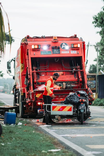 A street scene showing a sanitation worker standing beside a large red rubbish collection vehicle with the back open, revealing the internal mechanical components and loading mechanism. The worker, wearing a yellow reflective vest and dark trousers, appears to be managing or inspecting waste disposal, with a black trash bag placed on a small metal platform attached to the vehicle. The surrounding environment includes a sidewalk with a patch of grass featuring a blue container, and a background with trees, power lines, and a commercial building or shop partially visible on the right side. The scene is set during daylight hours with natural lighting and an overcast sky, emphasizing the practical aspect of private waste collection services distinct from municipal rubbish disposal. The composition highlights the process of on-site rubbish handling, illustrating the type of equipment used in independent waste removal activities, relevant to the scope of [PAGE_TITLE] and the broader context of alternative waste management solutions.