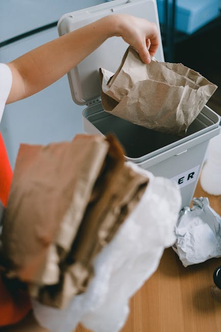 A person is seen disposing of a brown paper bag filled with rubbish into a white waste bin with a black liner, positioned on a wooden surface indoors. The person's arm is extended, holding the bag by its top edge, while the other hand is holding an additional paper bag in the foreground. The waste bin is placed on a table or countertop, with surrounding objects including crumpled paper and a small container nearby. The environment appears to be a domestic or office setting, and the scene relates to private waste disposal or rubbish collection services as part of alternative rubbish clearance options. The materials in the bags are made of brown paper, with some visible creasing and folds, and the bin has a smooth, matte finish. The lighting is neutral and evenly distributed, highlighting the practical nature of rubbish removal activities, which are a core element of private waste management services provided by companies like rubbishclearancewestkensington.com.