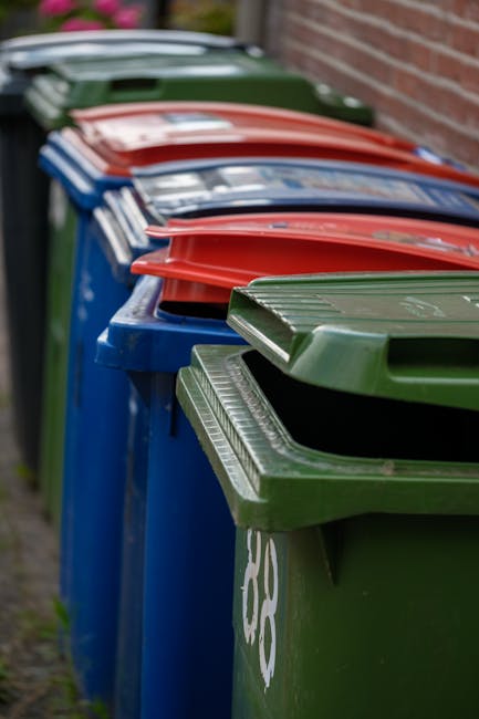A row of four large wheelie bins placed on a paved pathway alongside a brick wall, each with a different colored lid—green, blue, red, and another red—indicating separate waste or recycling compartments. The green bin at the front has a textured, ribbed lid with a slightly raised handle, and the blue bin behind it shows a faint recycling symbol on its side. The red-lidded bins further back are partially visible, with their lids closed and overlapping slightly in the line. The bins are situated outdoors, with the atmosphere suggesting an everyday rubbish collection area, possibly in a residential setting. Natural daylight illuminates the scene, casting soft shadows on the bins and pavement. This image reflects the common practice of local authority or private waste management for household rubbish, with colour-coded bins facilitating waste separation and pickup, consistent with alternative waste handling options outside of standard council collection services. The scene emphasizes the importance of organised waste disposal infrastructure in maintaining cleanliness and environmental responsibility in urban or suburban areas.
