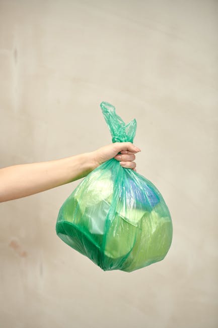 A person’s arm extends into the frame from the left, holding a green plastic bag tied at the top with a knot, containing garbage or waste material. The bag appears to be partially transparent with some visibility of contents inside, and has a smooth, slightly glossy surface. The background features a plain, neutral-colored wall with minimal texture, creating a neutral setting that emphasizes the bag and hand. The lighting is soft and even, ensuring details of the bag’s material and the arm are clearly visible. This scene may relate to waste disposal or rubbish collection, reflecting the context of independent or private rubbish handling services offered by companies like rubbishclearancewestkensington.com, aligning with themes of rubbish removal and waste management.