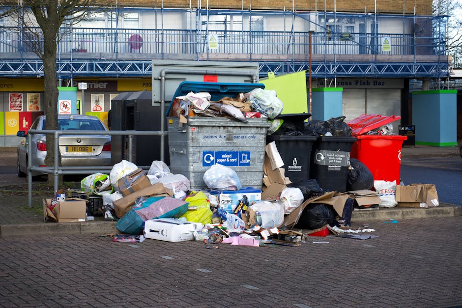 A row of four large wheelie bins placed on a paved pathway alongside a brick wall, each with a different colored lid—green, blue, red, and another red—indicating separate waste or recycling compartments. The green bin at the front has a textured, ribbed lid with a slightly raised handle, and the blue bin behind it shows a faint recycling symbol on its side. The red-lidded bins further back are partially visible, with their lids closed and overlapping slightly in the line. The bins are situated outdoors, with the atmosphere suggesting an everyday rubbish collection area, possibly in a residential setting. Natural daylight illuminates the scene, casting soft shadows on the bins and pavement. This image reflects the common practice of local authority or private waste management for household rubbish, with colour-coded bins facilitating waste separation and pickup, consistent with alternative waste handling options outside of standard council collection services. The scene emphasizes the importance of organised waste disposal infrastructure in maintaining cleanliness and environmental responsibility in urban or suburban areas.