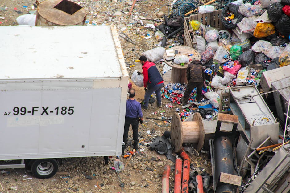 Aerial view of a large area filled with mixed waste and debris, including various plastic bags, cardboard, and household items. In the center, three individuals are actively engaged in sorting or collecting rubbish, with one person wearing a red and black jacket, another in dark clothing, and a third in a purple top. To the left, a white commercial truck with the registration number 'AZ 99-FX-185' is parked, partially blocking the scene. The ground is uneven with dirt and smaller debris scattered around. Among the waste, there are large wooden spools, metal pipes, and discarded household appliances, creating a cluttered environment characteristic of illegal dumping sites or construction debris. The waste pile extends to the upper right corner of the image, where more bags and scattered refuse are visible. The outdoor setting appears poorly maintained, with no clear organization, highlighting issues related to private waste removal or unmanaged rubbish accumulation, relevant to independent rubbish clearance services.