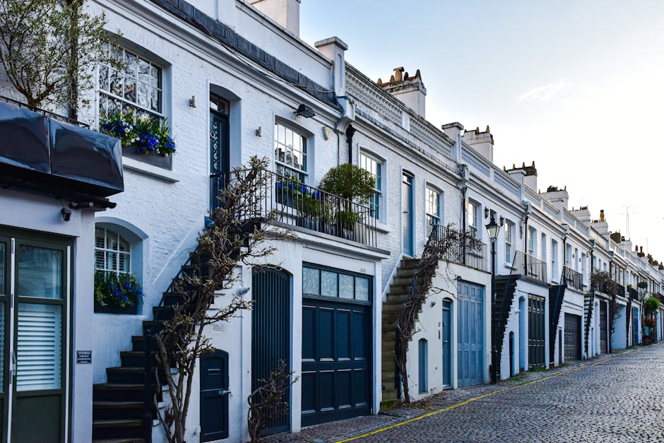 The image depicts a row of white terraced residential buildings with blue accents, located along a cobbled street. Each property features a small front garden or planter box, some containing potted plants with green foliage and colorful flowers. External staircases with black iron railings lead up to individual entrances, which are painted in shades of blue, matching the garage doors on the ground level. The texture of the building exteriors appears to be roughcast or brick, painted white, with some windows adorned with decorative window boxes filled with vibrant flowers. The street is empty, with natural daylight illuminating the scene, casting soft shadows. The buildings are closely spaced, with minimal space between them, and the lack of visible rubbish or clutter suggests a well-maintained area, possibly within a conservation or residential zone. Occasional details such as a lamppost and a small tree leaning near a staircase contribute to the suburban atmosphere. The scene aligns with contexts where local authorities or private services might handle refuse outside such properties, especially in historical or affluent neighbourhoods, supporting the theme of private waste handling and alternative rubbish removal access issues.