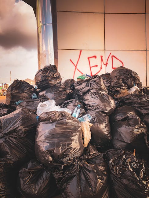 Aerial view of a large area filled with mixed waste and debris, including various plastic bags, cardboard, and household items. In the center, three individuals are actively engaged in sorting or collecting rubbish, with one person wearing a red and black jacket, another in dark clothing, and a third in a purple top. To the left, a white commercial truck with the registration number 'AZ 99-FX-185' is parked, partially blocking the scene. The ground is uneven with dirt and smaller debris scattered around. Among the waste, there are large wooden spools, metal pipes, and discarded household appliances, creating a cluttered environment characteristic of illegal dumping sites or construction debris. The waste pile extends to the upper right corner of the image, where more bags and scattered refuse are visible. The outdoor setting appears poorly maintained, with no clear organization, highlighting issues related to private waste removal or unmanaged rubbish accumulation, relevant to independent rubbish clearance services.